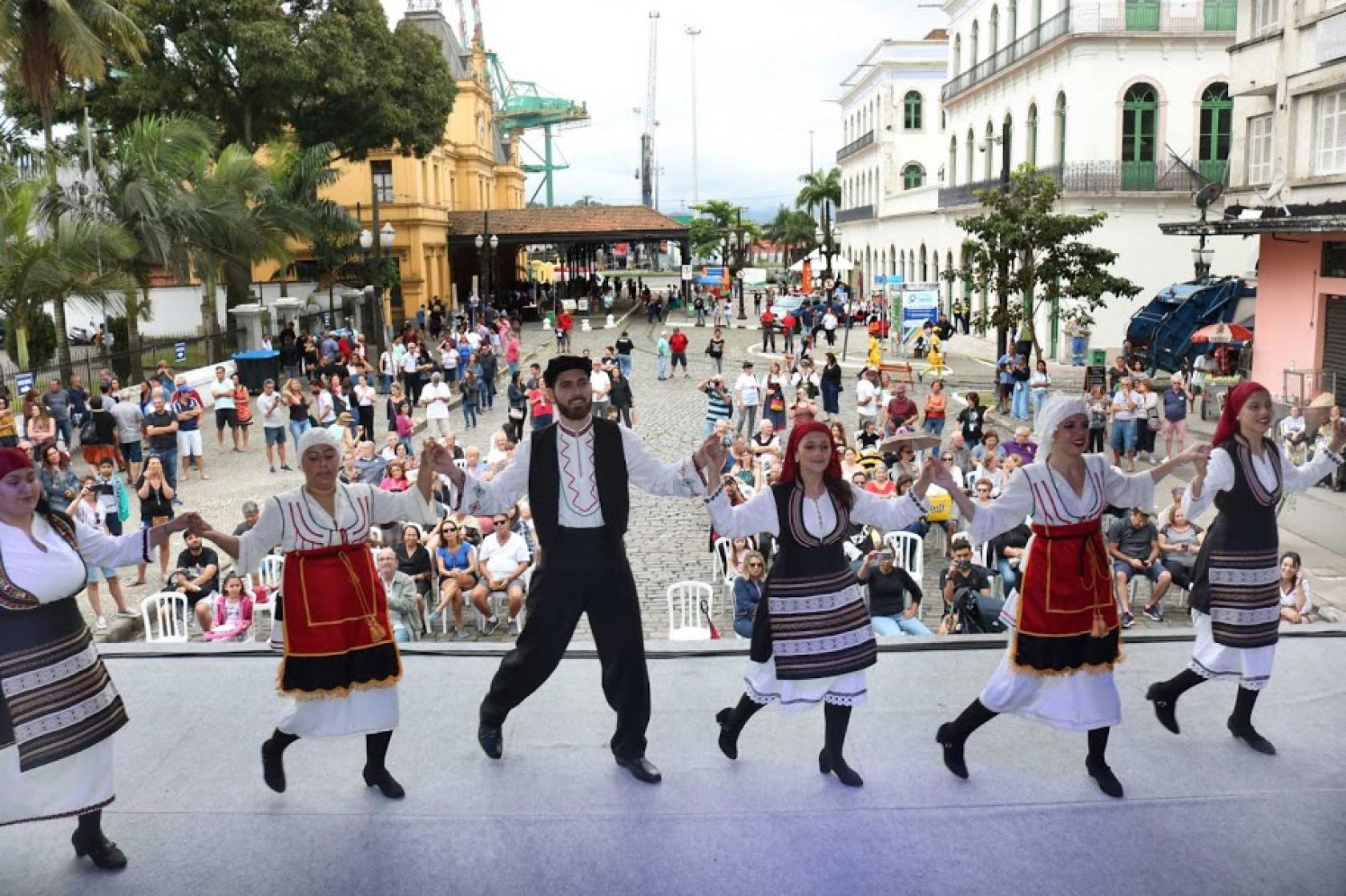 Dançarinos típicos se apresentam de costas para o público com Museu Pelé e Estação do Valongo ao fundo. #pracegover