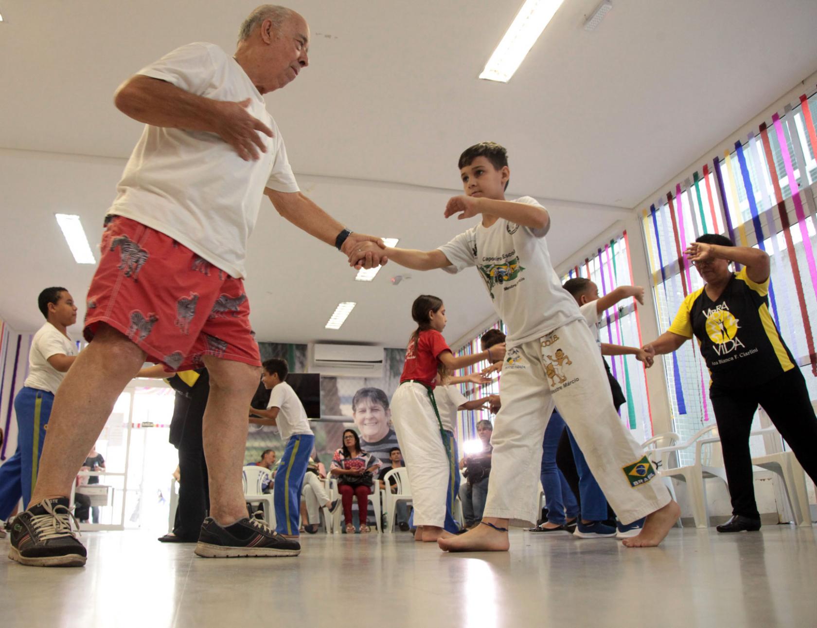 Idoso e menino jogam capeira em salão onde outras duplas também jogam. #Pracegover