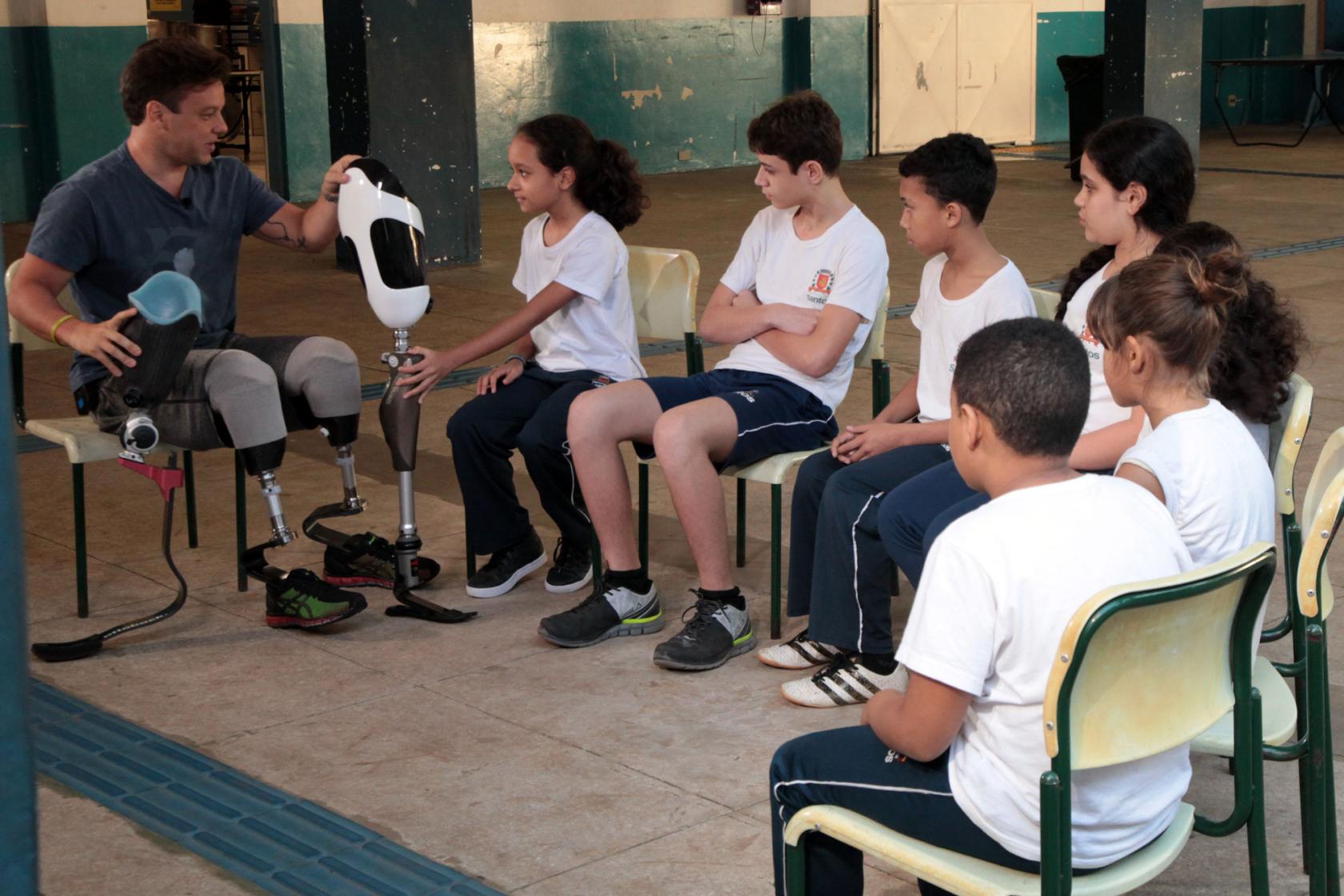 Pauê está sentado em uma cadeira usando e mostra exemplar de próteses para os alunos. Uma das meninas, ao lado dele, toca na peça. Os outros estudantes observam. #Pracegover