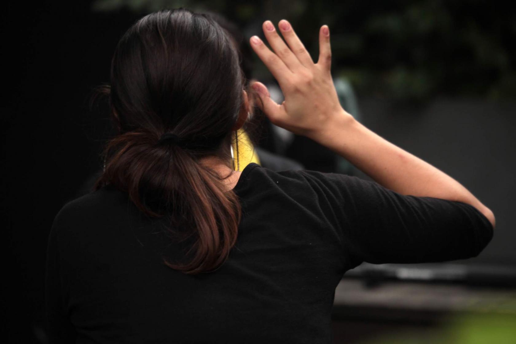 Mulher está de costas para a foto. Ela tem os cabelos lisos e compridos. Seu braço direito está erguido até a altura da cabeça. A mão está aberta, como num sinal de Pare. #Pracegover