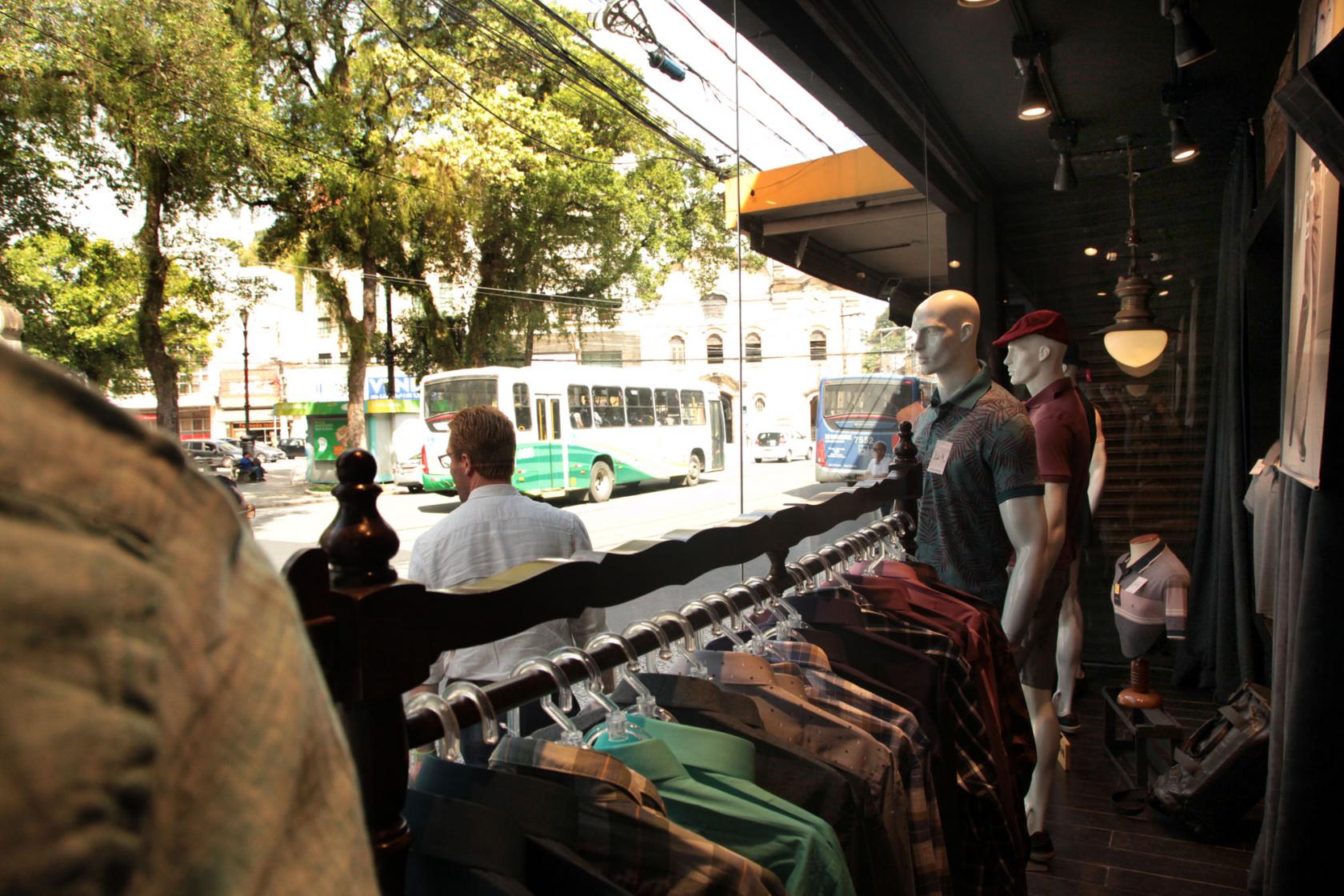 Imagem feita de dentro de loja com vista para uma praça. Dentro da loja estão camisas penduradas em araras e manequins com roupas. Na rua passa uma pessoa e um ônibus ao fundo. #Pracegover