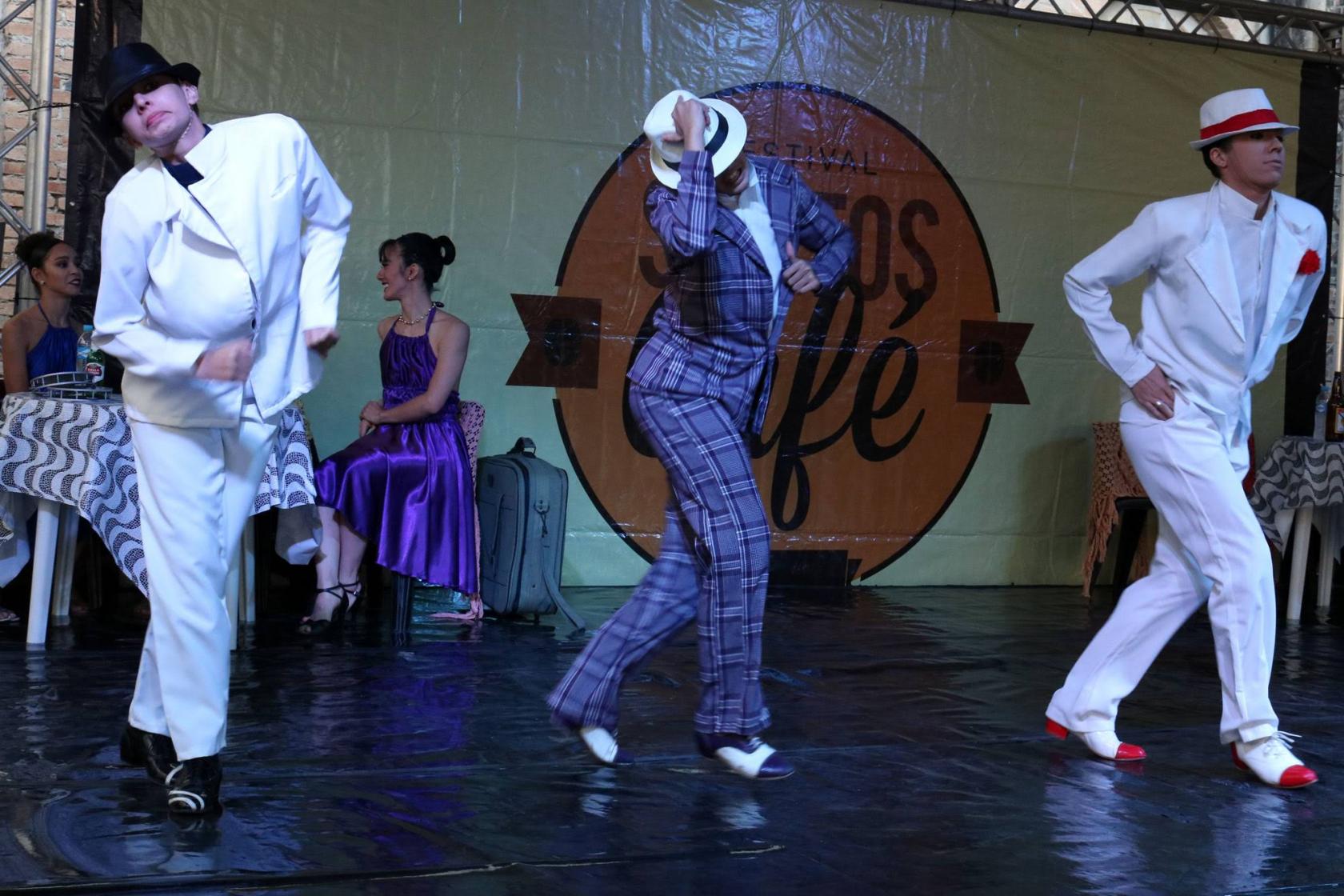 Balé da Cidade apresenta coreografia. Há três bailarinos no palco. Eles usam roupas masculinas e chapéus. #Pracegover