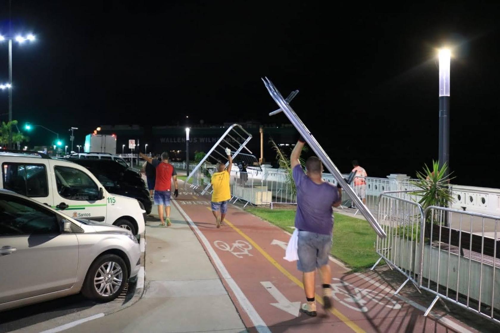 homens carregam gradis para serem instalados no acesso ao calçadão, na ponta da Praia. Alguns já estão instalados. É noite e os postes estão acesos. #Paratodosverem