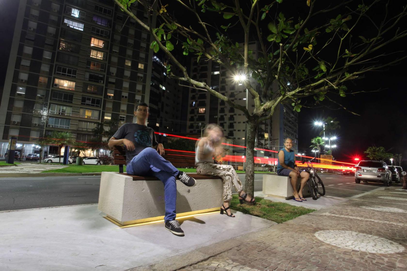 Pessoas sentadas em banco iluminado #pracegover