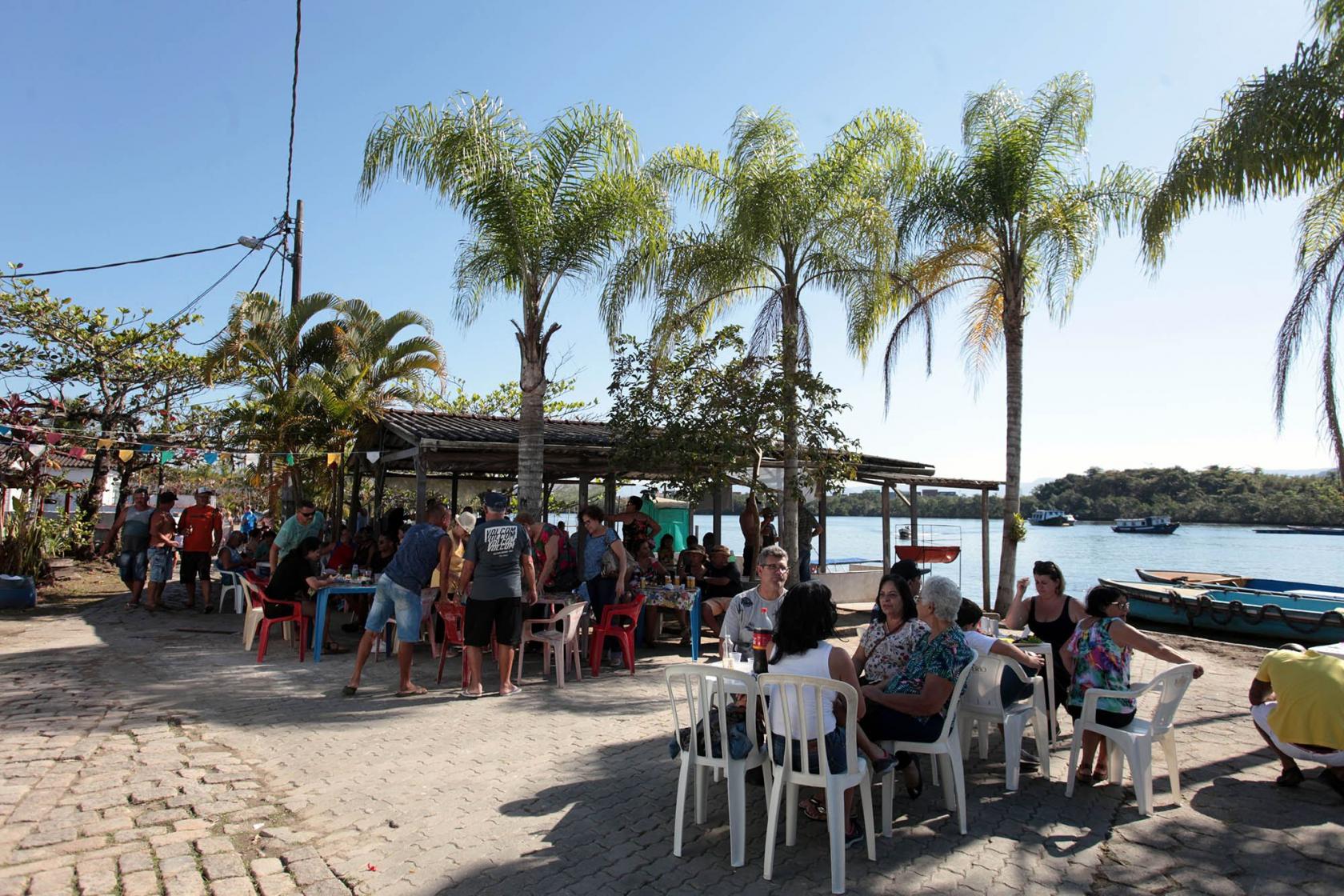 Cerca de 50 pessoas em pé e sentadas junto a mesas comem e bebem em área aberta com palmeiras junto ao mar #pracegover