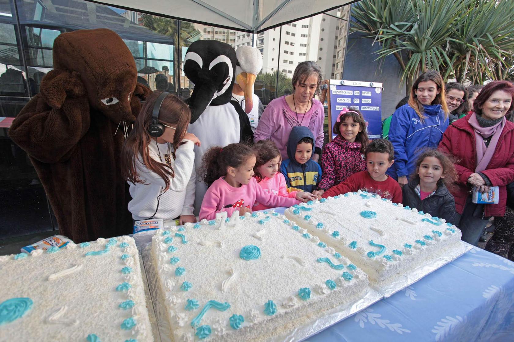 Crianças cantam parabéns diante de bolo e acompanhadas por grandes bonecos de animais marinhos. #Pracegover