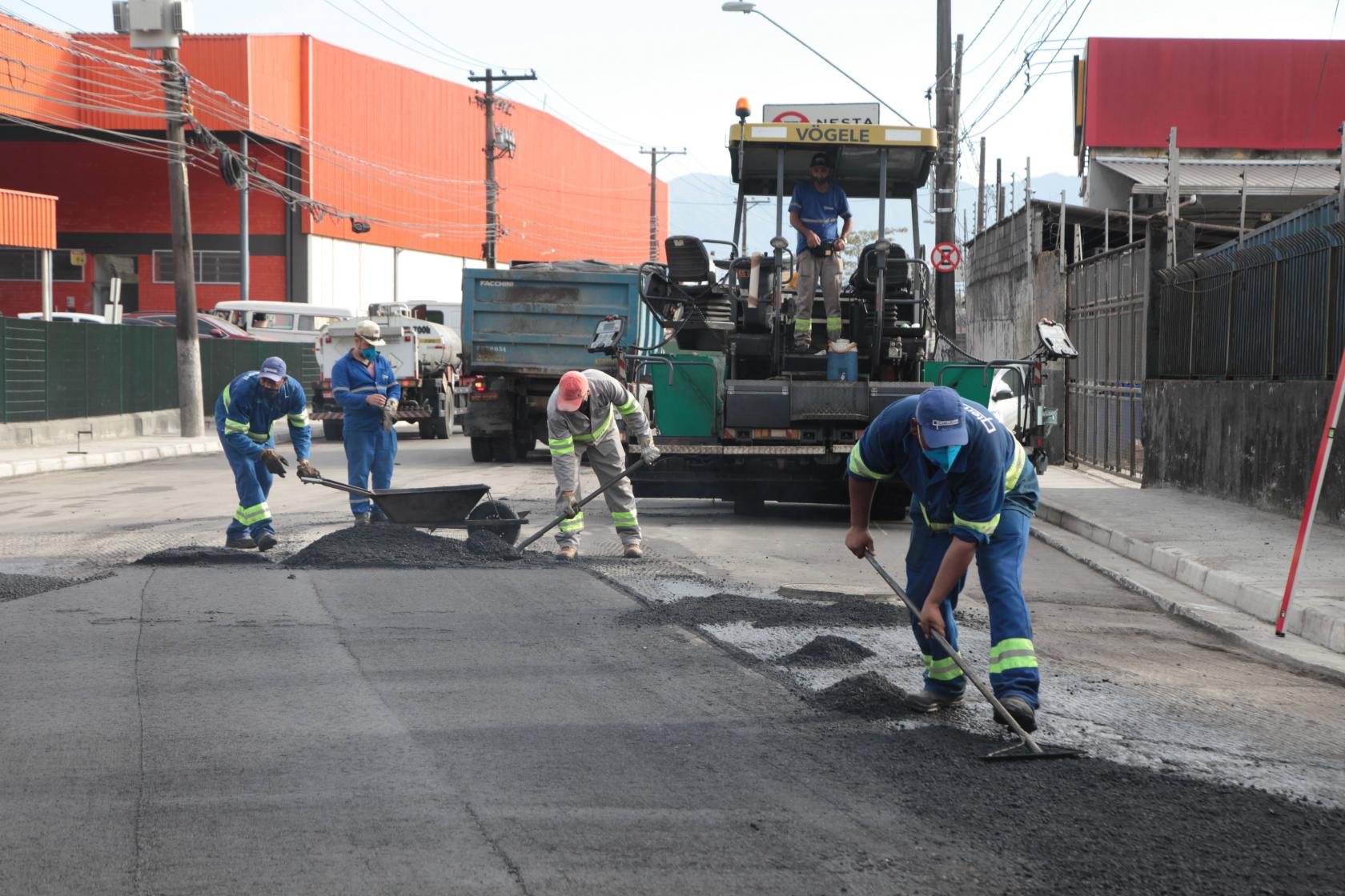 Trabalhadores de uniforme azul com enchadas na mão espalhando piche pela rua no trabalho de recapeamento