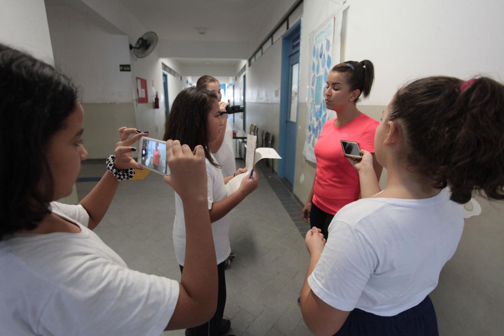 Alunos filmam, fotogravam e entrevistam uma mulher no corredor de escola. #Pracegover