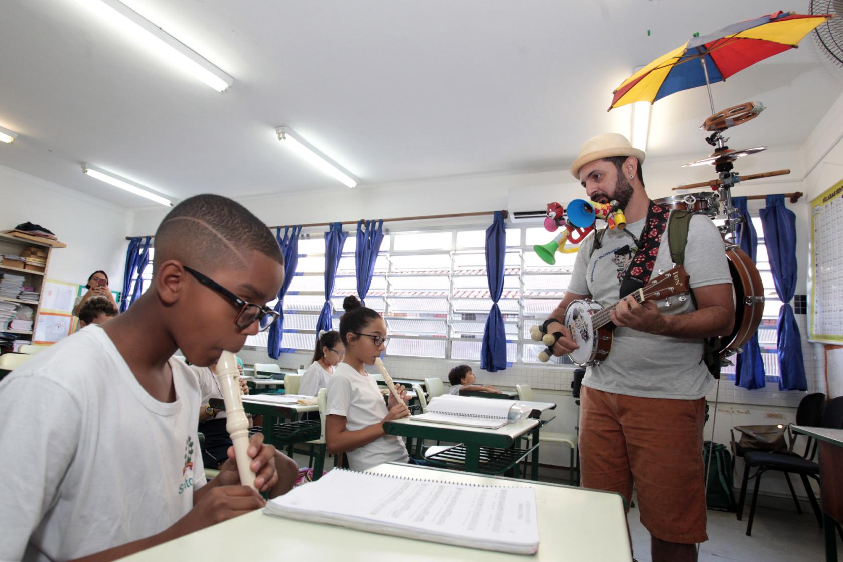 músico está em pé, com guarda chuva nas costas, tocando com aluno que está sentando tocando flauta #pracegover 