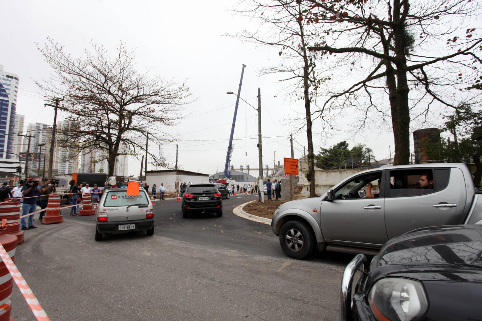 Fluxo de veículos em trecho de via. Há árvores e postes. A pista foi recém-asfaltada. #Pracegover