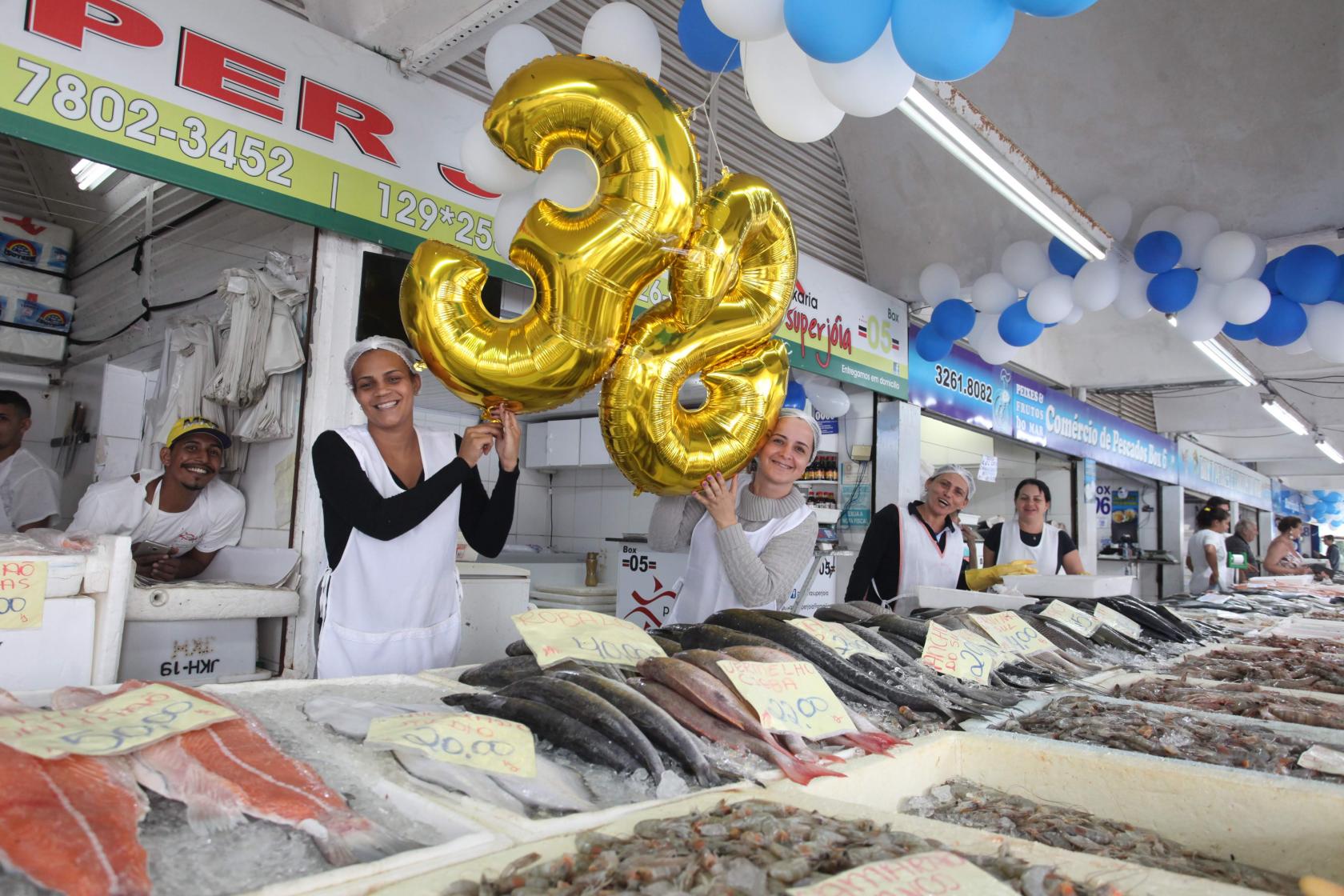 funcionárias seguram balões formando número 38 ao lado de peixes à venda #pracegover 