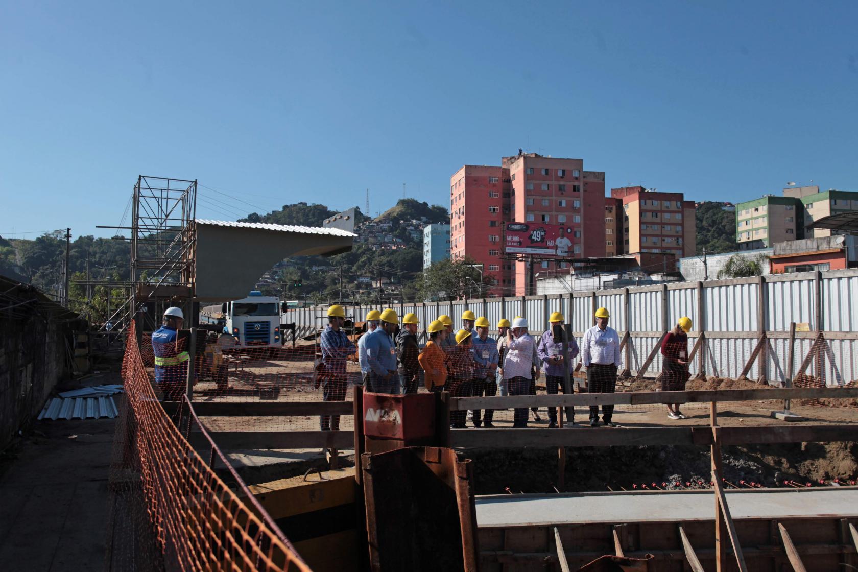 Obras na entrada da Cidade, com conjunto habitacional ao fundo. Pessoas circulam pela obra. #Pracegover