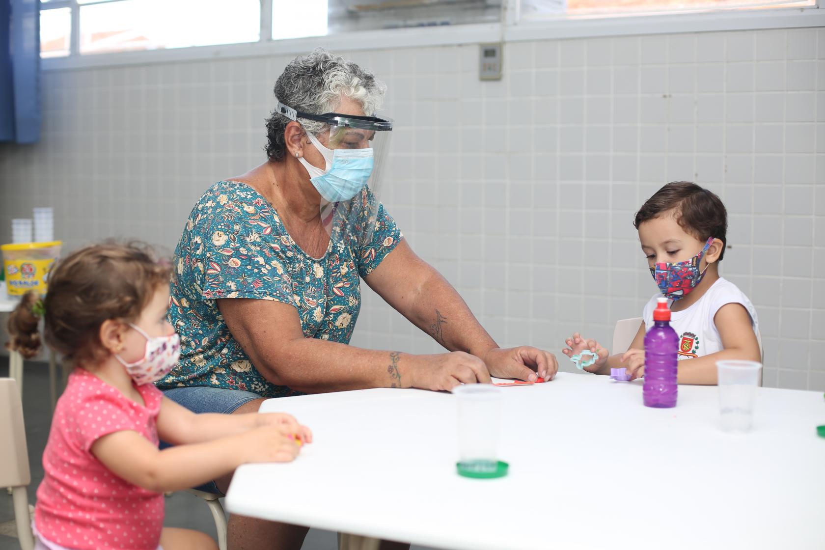 professora brinca em mesa com dois alunos #paratodosverem