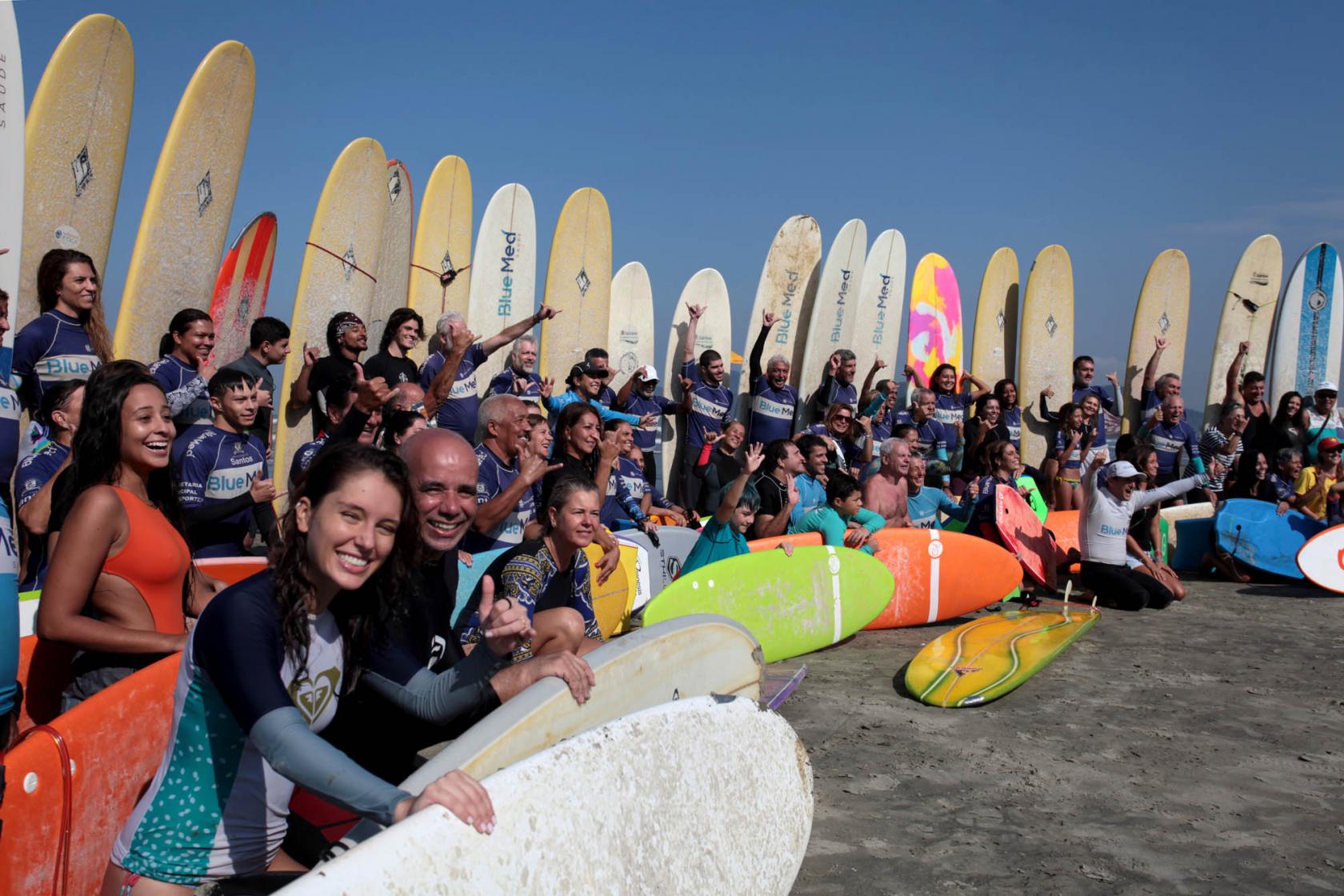 Dezenas de surfistas postam para fotos. Ao fundo há um paredão de pranchas. Os atletas estão na frente. Os que estão na fileira da frente estão com as pranchas sobre a areia na transversal. #Pracegover