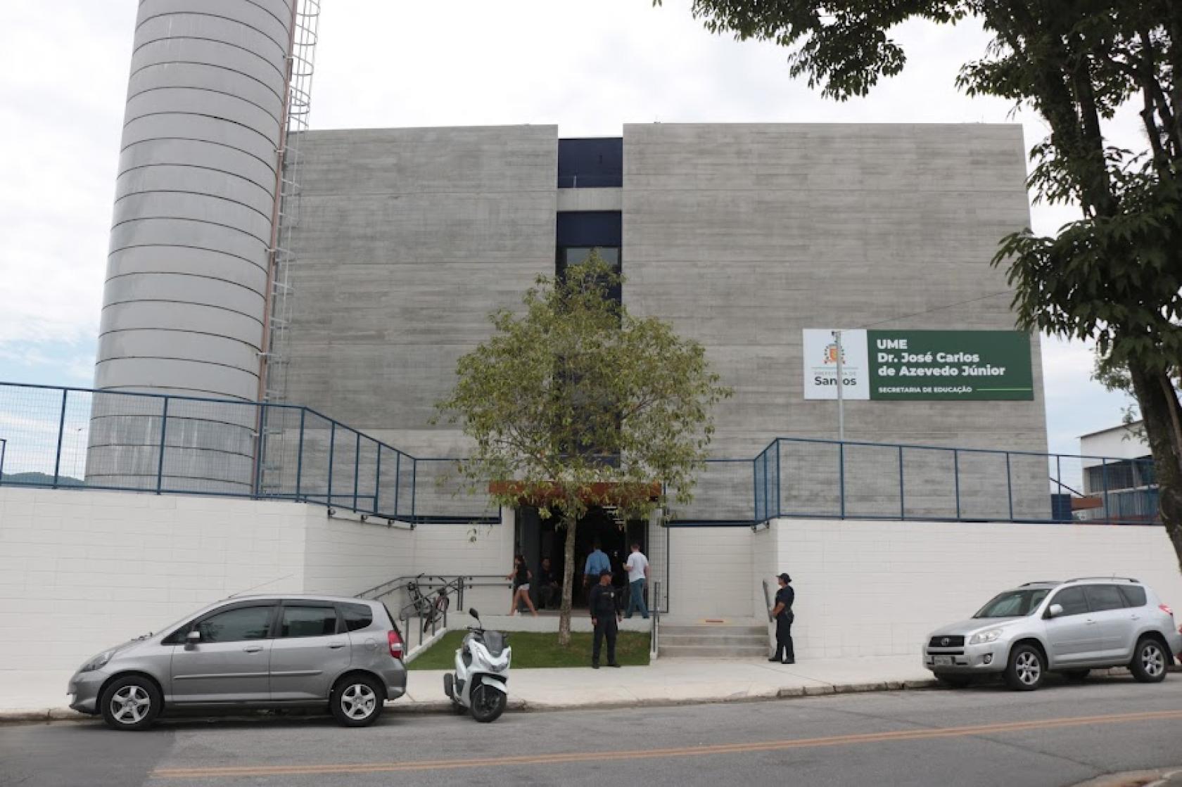 fachada da escola com árvore na entrada. há dois carros e uma mota estacionados na frente. A escola tem um placa de identificação. #paratodosverem