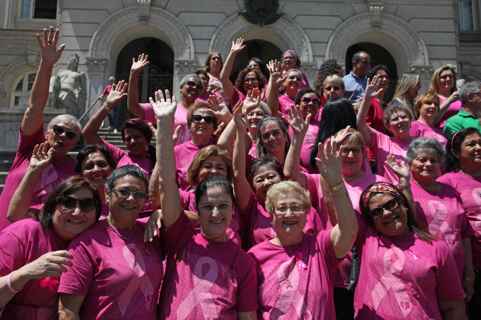 Mulheres vestidas com camisetas cor de rosa posam para foto em frente à Prefeitura. Elas estão com os braços para o alto.#Pracegover