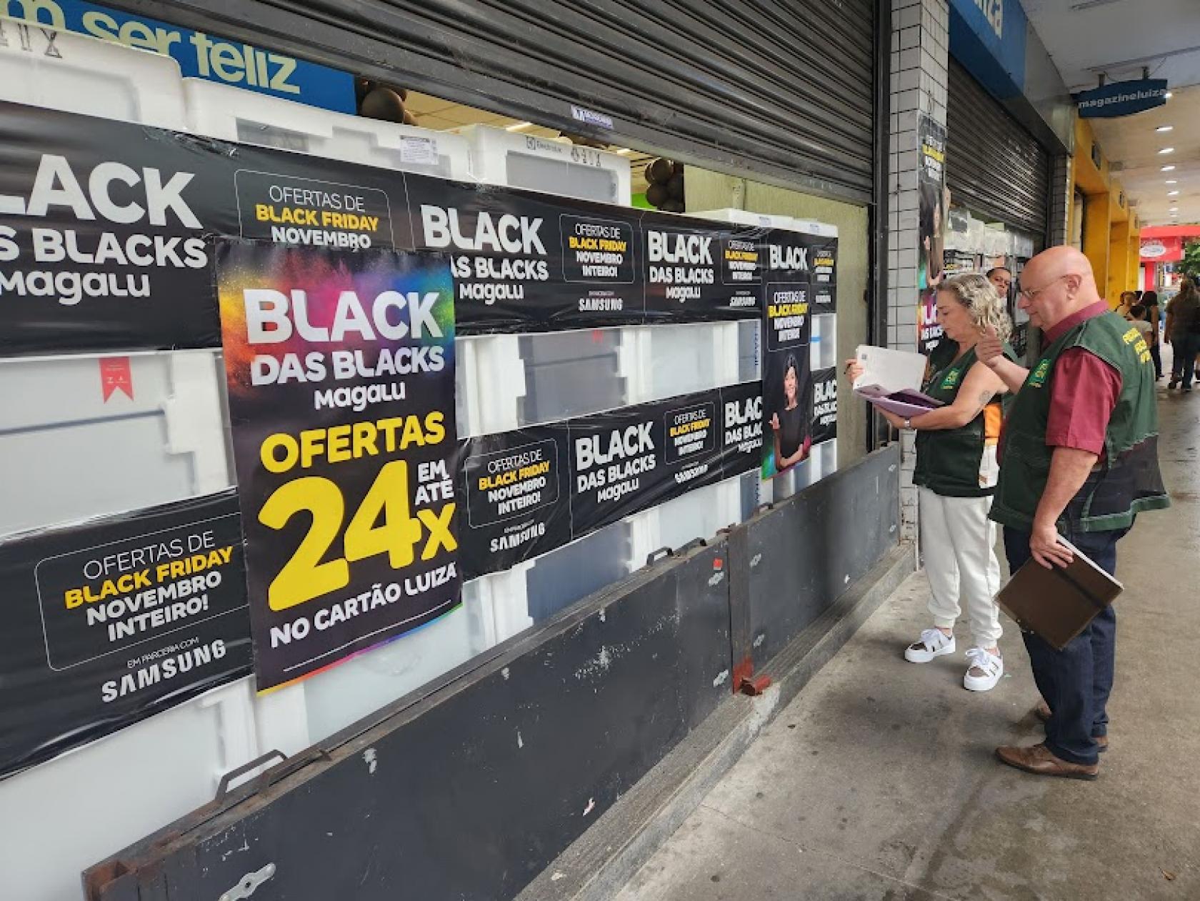 dois agentes uniformizados do procon estão na rua diante da vitrine de uma loja com cartazes sobre a black friday. #paratodosverem 