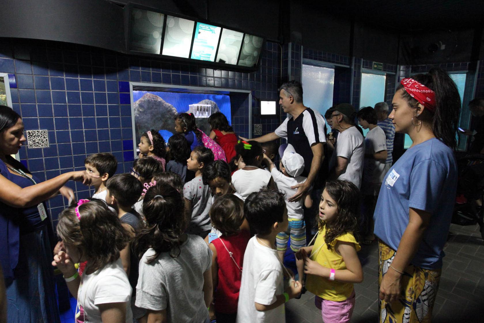 Muitas crianças estão passeando em corredor do Aquário. Elas estão conversando e observando os tanques. #Pracegover