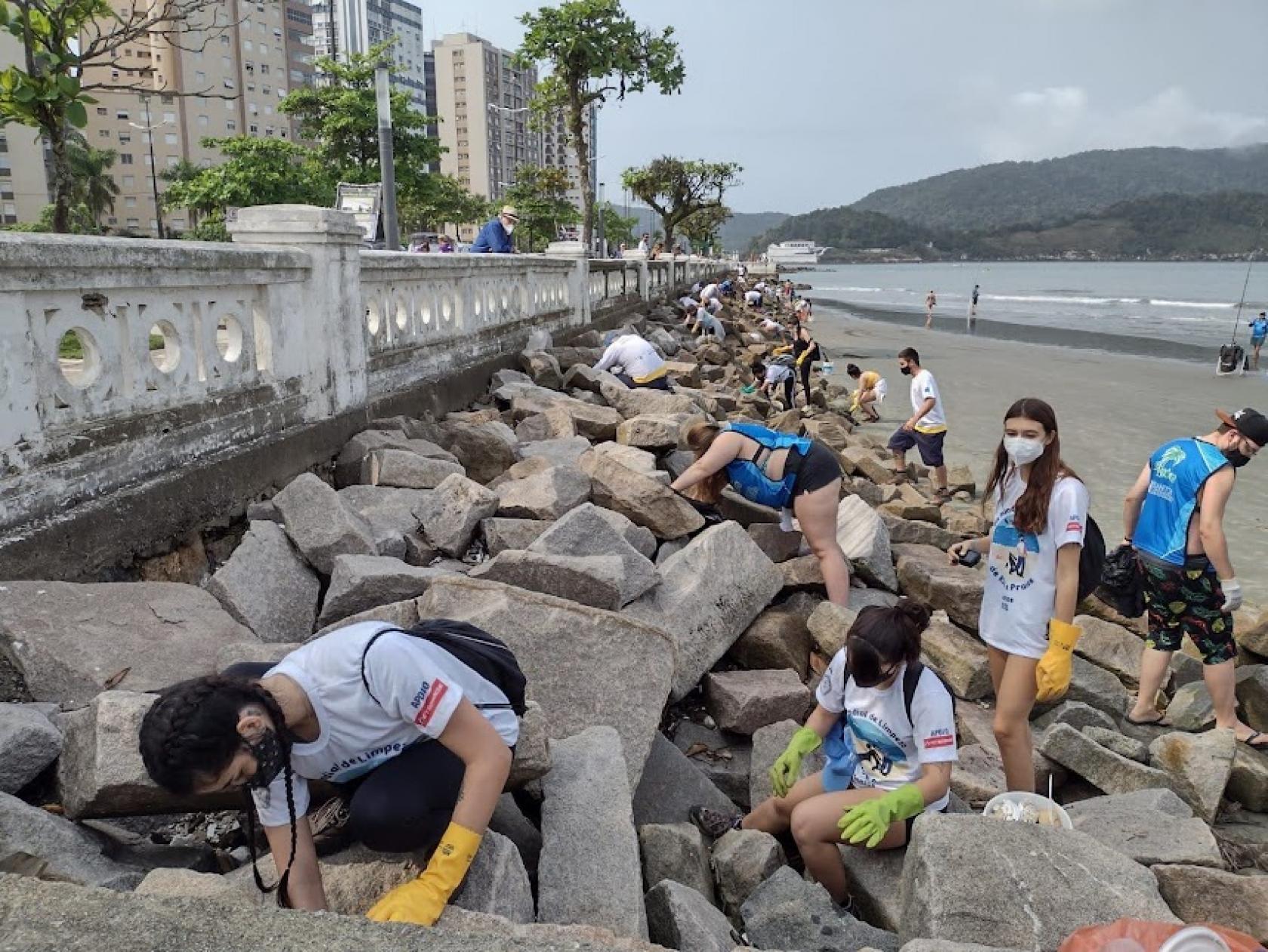 várias pessoas uniformizadas catando lixo no costão rochoso próximo à mureta na ponta da praia.Eles usam luvas e carregam sacos #paratodosverem