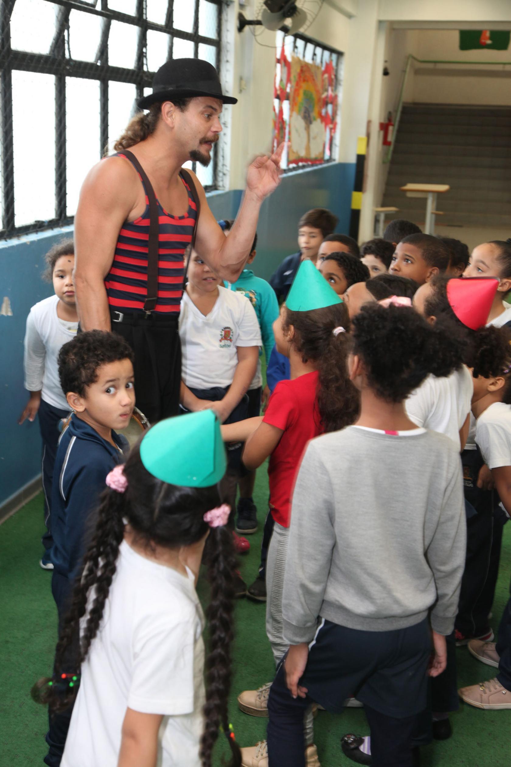 Ator vestido de palhaço está rodeado pelos alunos #pracegover