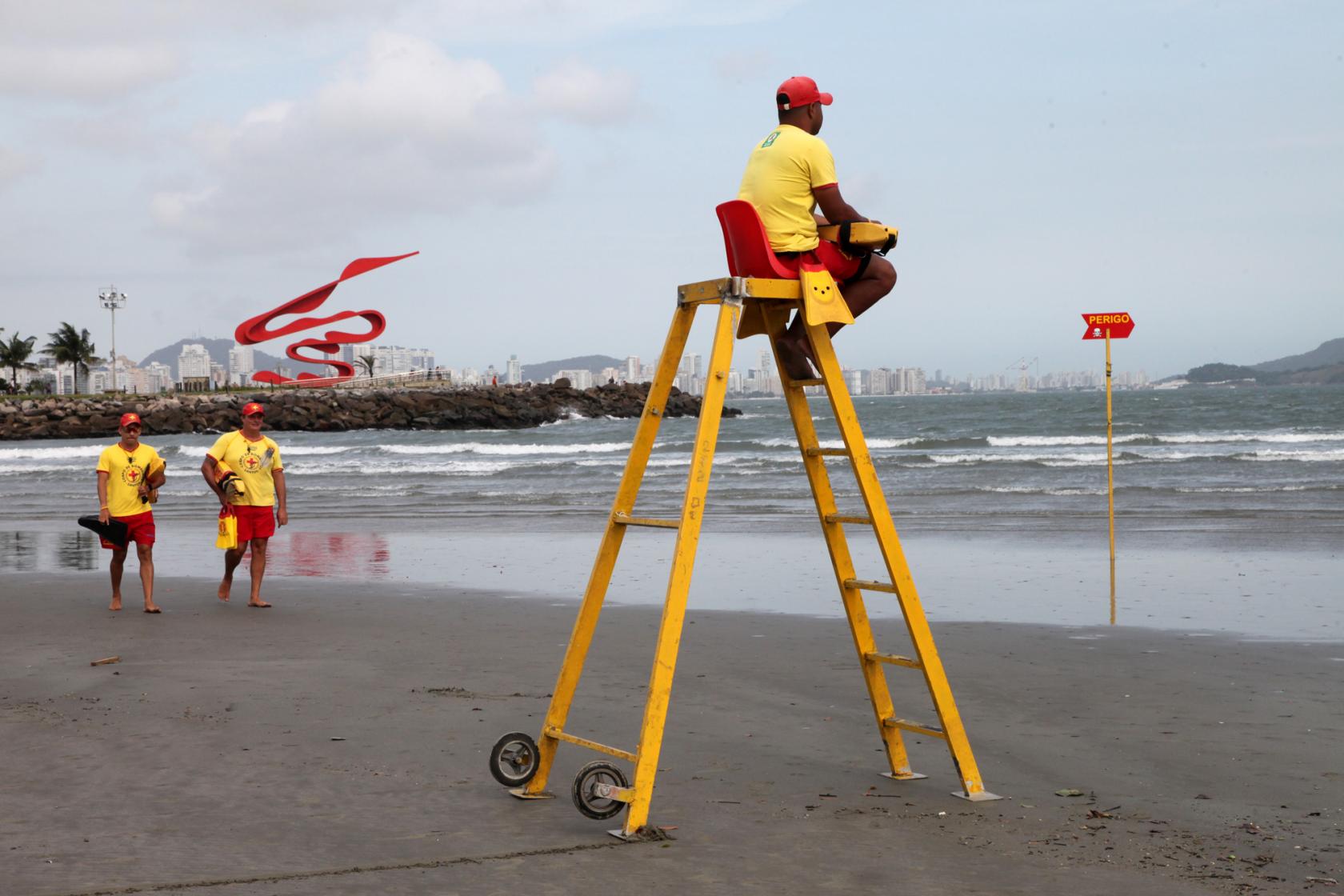 Em primeiro plano há uma escada armada na praia, com um salva vidas sentado no topo, onde há um banco. Ao fundo, outros dois guardas vêm andando. Eles estão de frente para o mar. #Pracegover