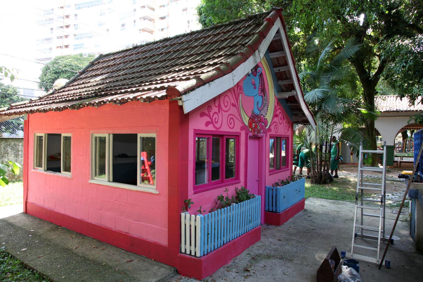 Casa de boneca com fachada frontal e lateral à vista. Lateral tem duas janelas abertas. Na frente, há duas janelas, porta e dois cercadinhos para plantas. Na frente há decoração com motivos infantis.  #Pracegover