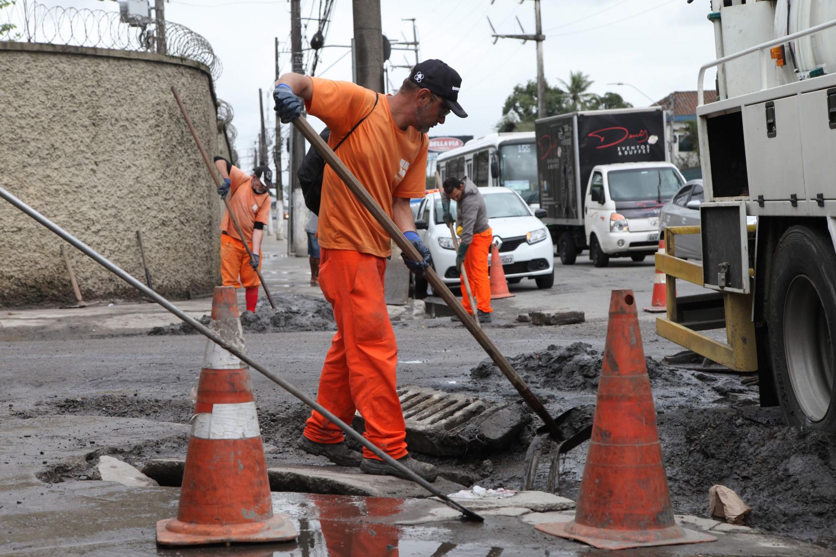 Homem remove lama de bueiro em rua. Há cones delimitando a área de liimpeza