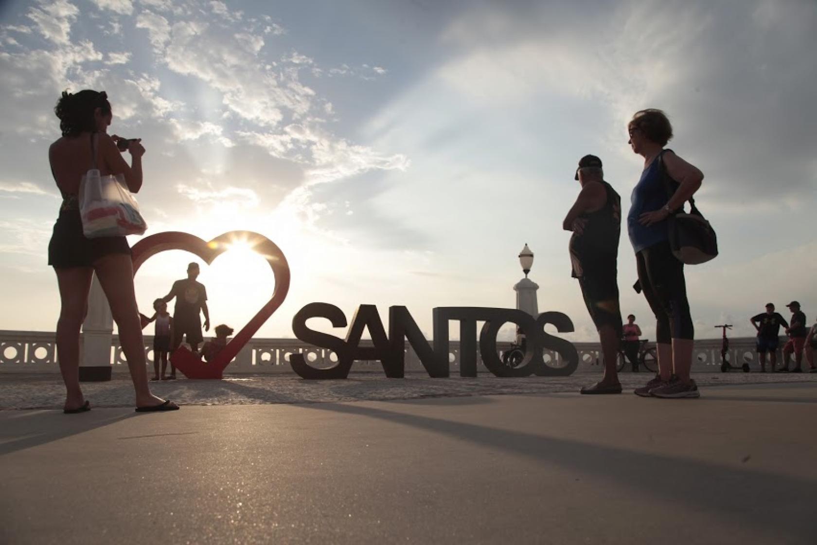 Monumento com a inscrição Santos e coração estilizado ao lado com pessoas olhando e tirando foto. #pracegover