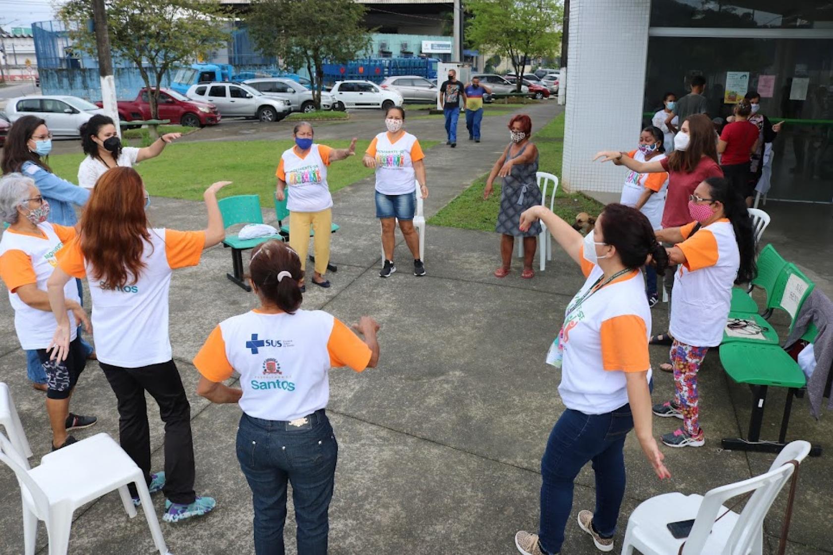 ATIVIDADE AO AR LIVRE. VÁRIAS PESSOAS ESTÃO EM PÉ, EM RODA, FAZENDO MOVIMENTOS COM OS BRAÇOS. #PARATODOSVEREM