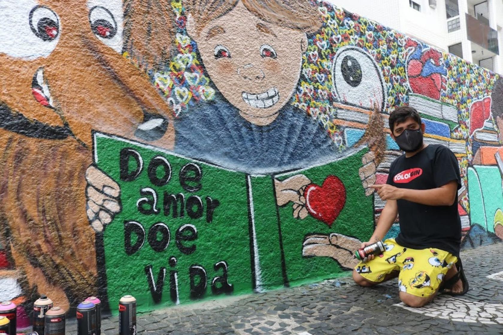 Mural grafitado com livro aberto onde se lê Doe Amor doe vida. O artista etá agachado no chão, segurando uma lata de tinta spray. #paratodosverem