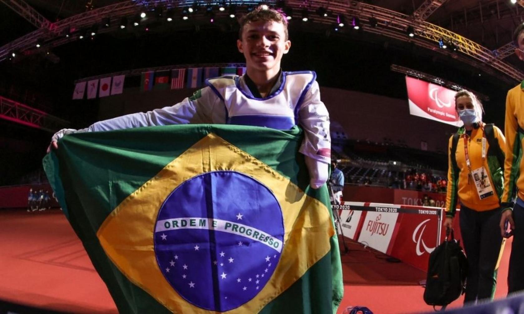 Nathan segura a bandeira do brasil na frente do corpo. Ele está dentro da arena. #paratodosverem