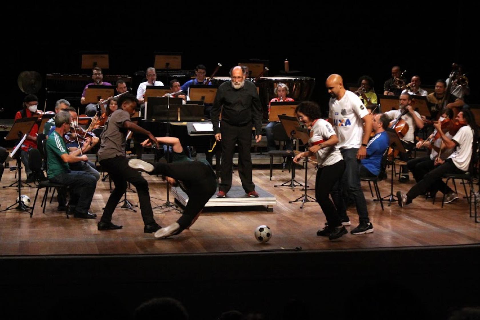 músicos da orquestra tocando e quatro pessoas encenando a disputa de um jogo de futebol no palco. #paratodosverem