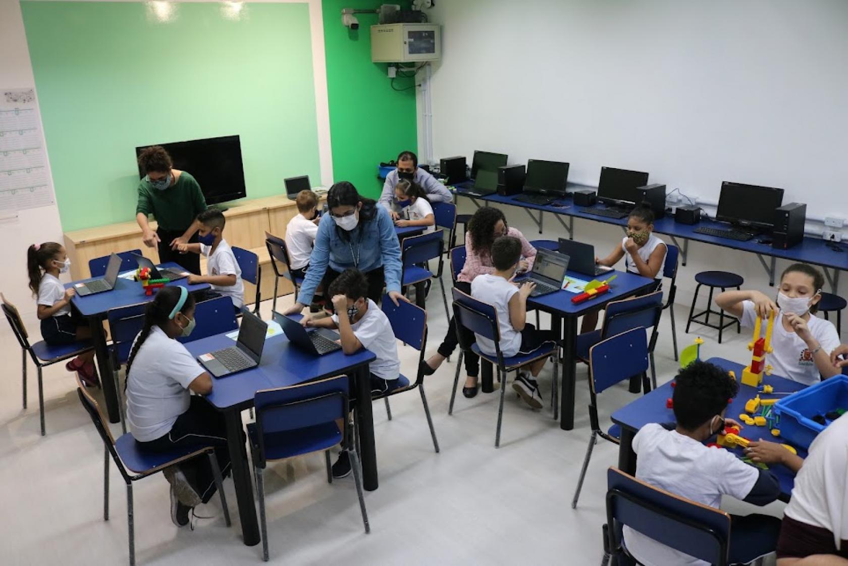 sala com diversas mesas. Alunos sentados mexendo nos computadores. Alguns professores estão ao lado dos alunos. À direita, uma menina monta uma estrutura com peças colorida, como um jogos de peças de encaixe. #paratodosverem