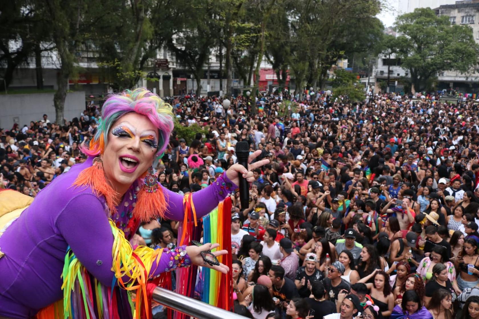 Drag Queen está no alto de um carro alegórico saudando uma multidão em praça. #Pracegover