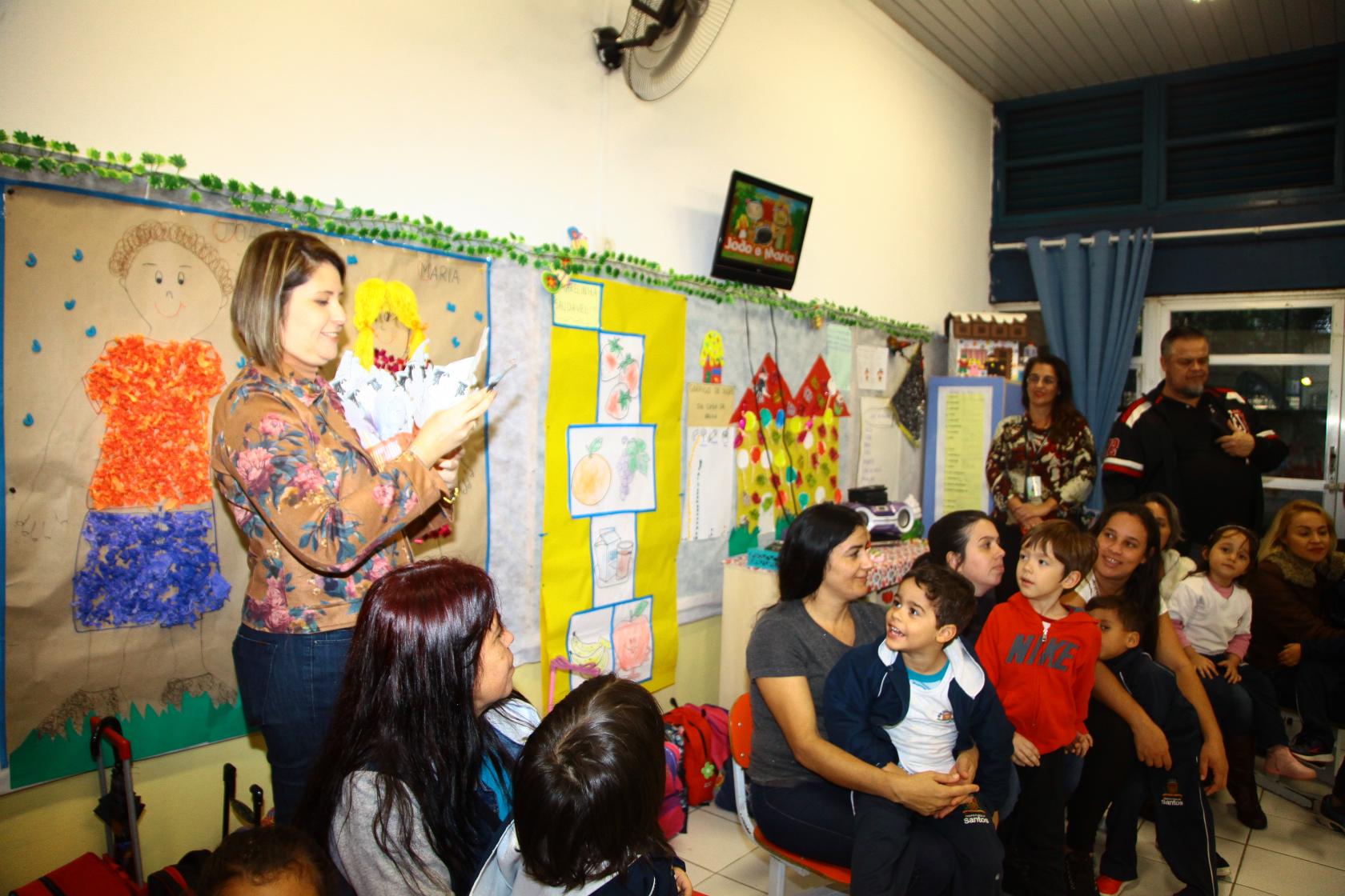 Professora, pais e alunos reunidos em uma sala de educação infantil. A educadora está fazendo uma apresentação aos pais. A parede da sala tem várias colagens de motivos infantis. #Pracegover