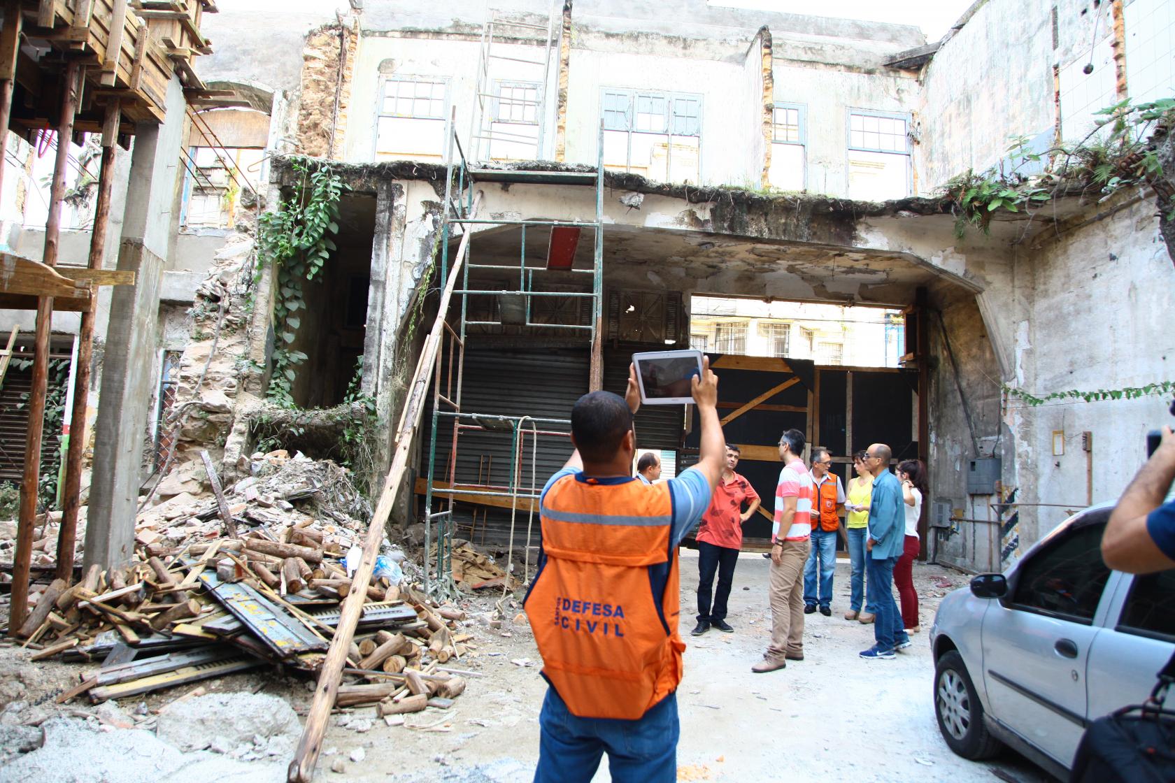 Agentes da Prefeitura estão na área interna de um imóvel parcialmente destruído. Um dos agentes, de costas para a imagem e com jaleco laranja está fotografando o espaço. #Pracegover