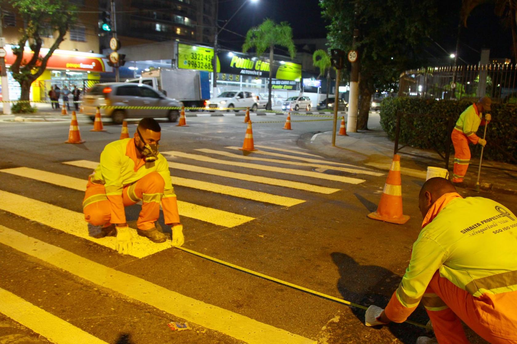 Homens demarcam faixa em avenida. Eles estão agachados s segurando uma especie de trena. Eles usam máscaras de proteção. Ao lado há cones demarcando a a´rea reservada para o serviço e para o trânsito de veículos. #Pracegover