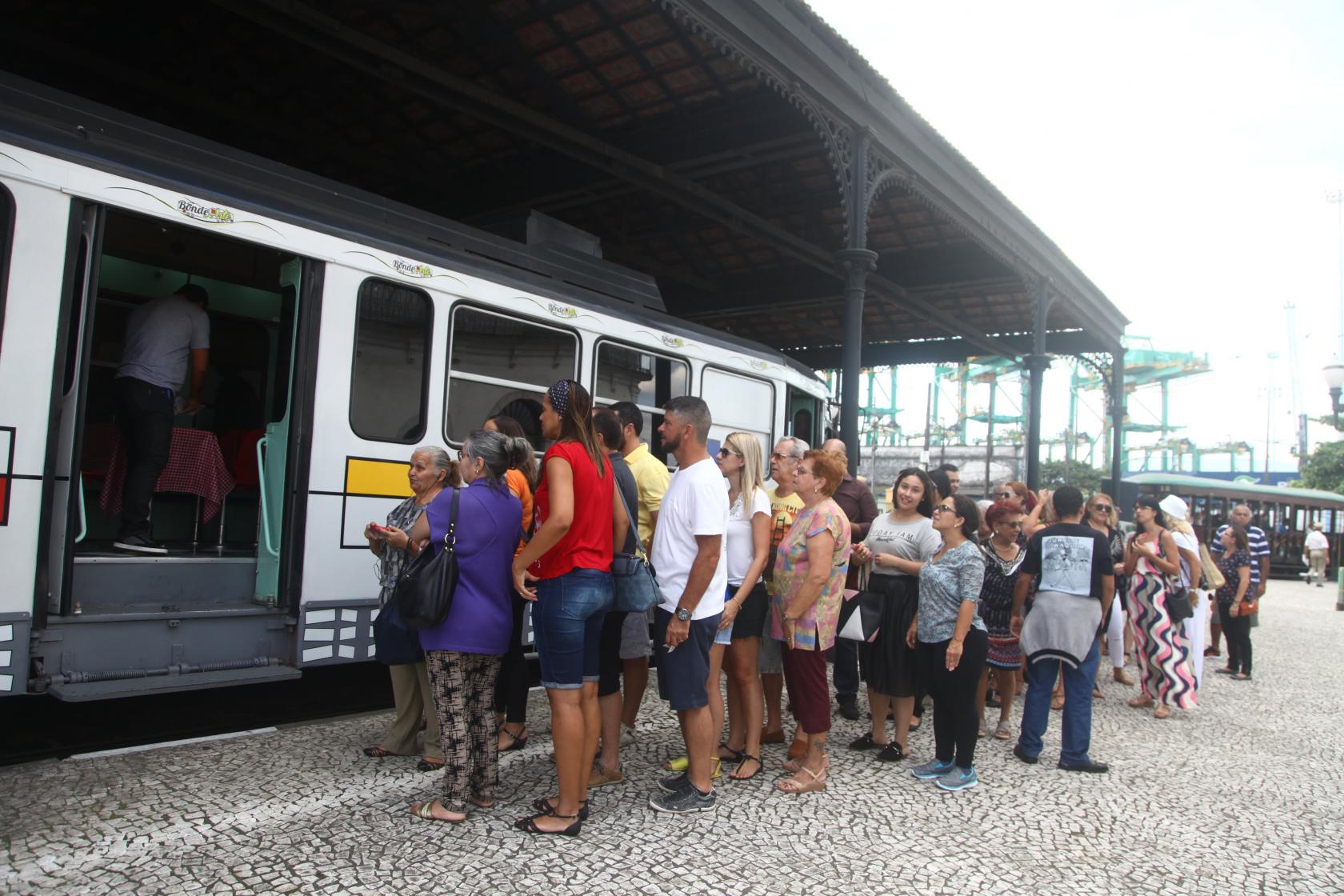 Pessoas fazem fila para ingressar no bonde arte. Ele está parado em estação. #Pracegover