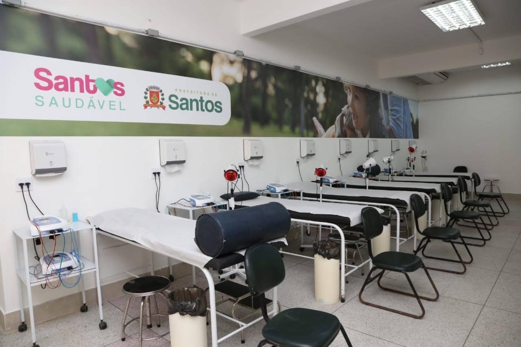 Sala com diversas macas de fisioterapia enfileiradas. Ao fundo há um painel onde se lê Santos Saudável. #Paratodosverem