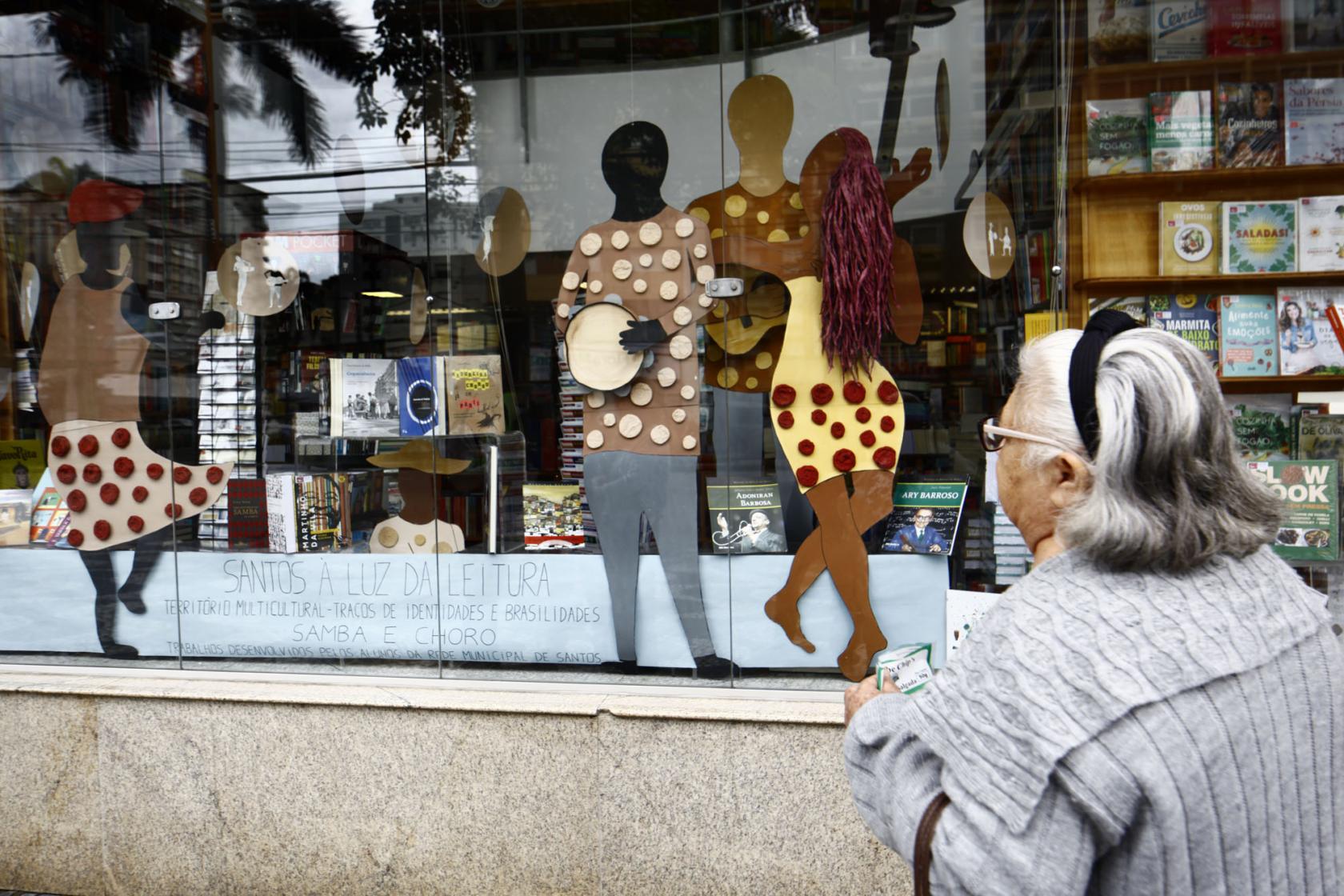 Figuras em papelão e coloridas expostas em vitrine. Uma mulher está olhando do lado de fora. #Pracegover