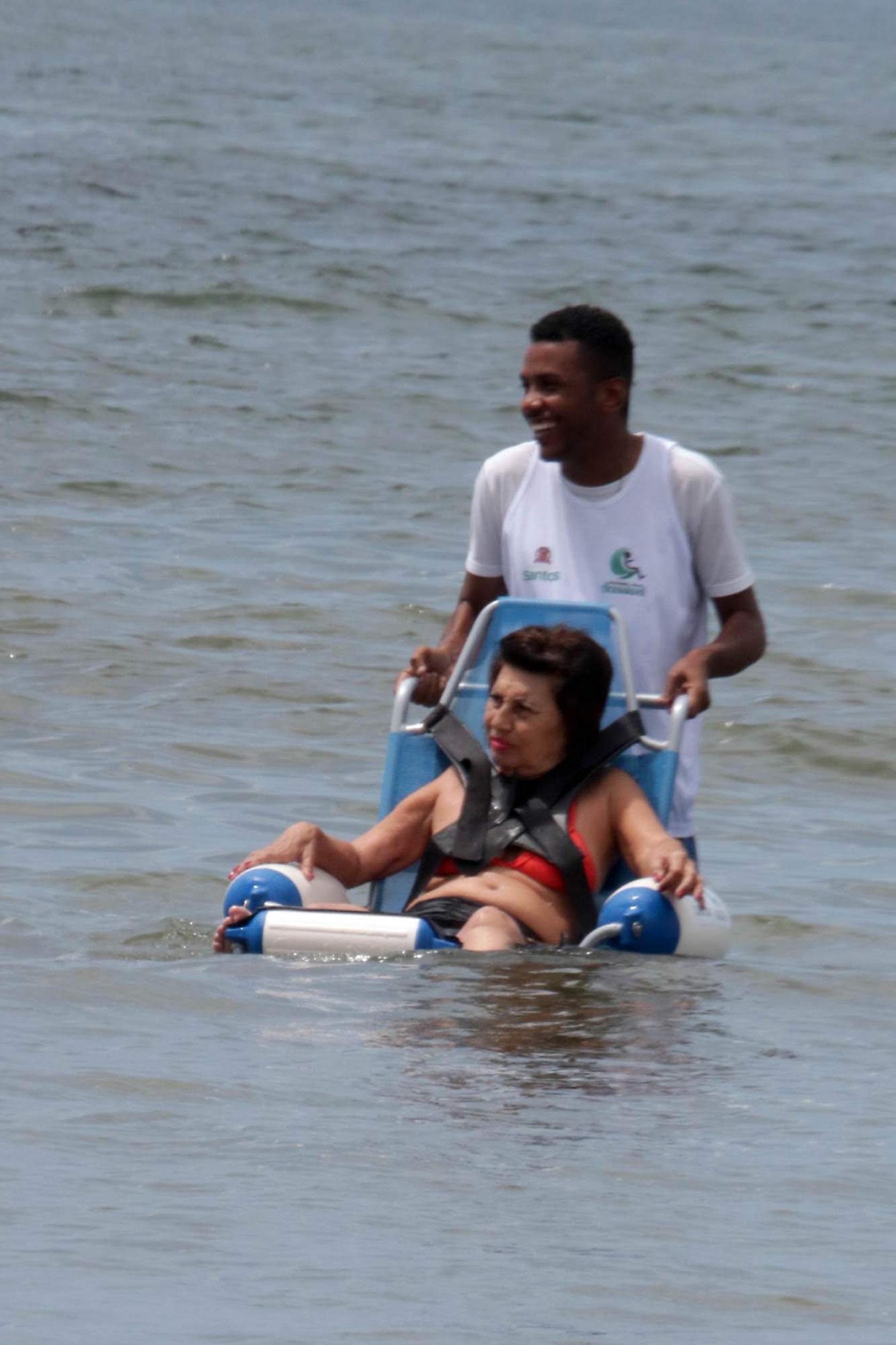 senhora toma banho de mar na cadeira ajudada por rapaz #pracegover