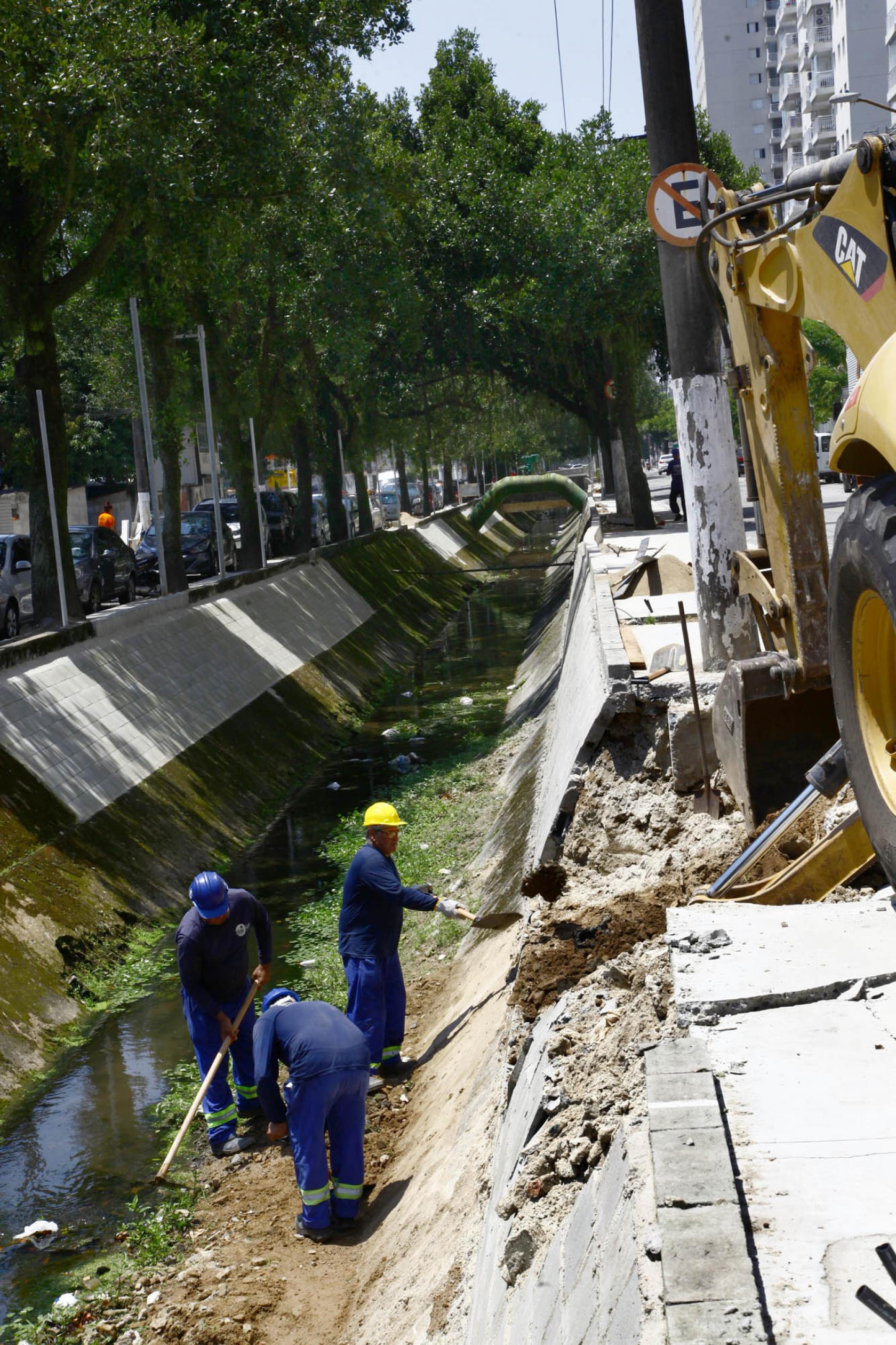 operários e máquina trabalham em borda de canal #pracegover