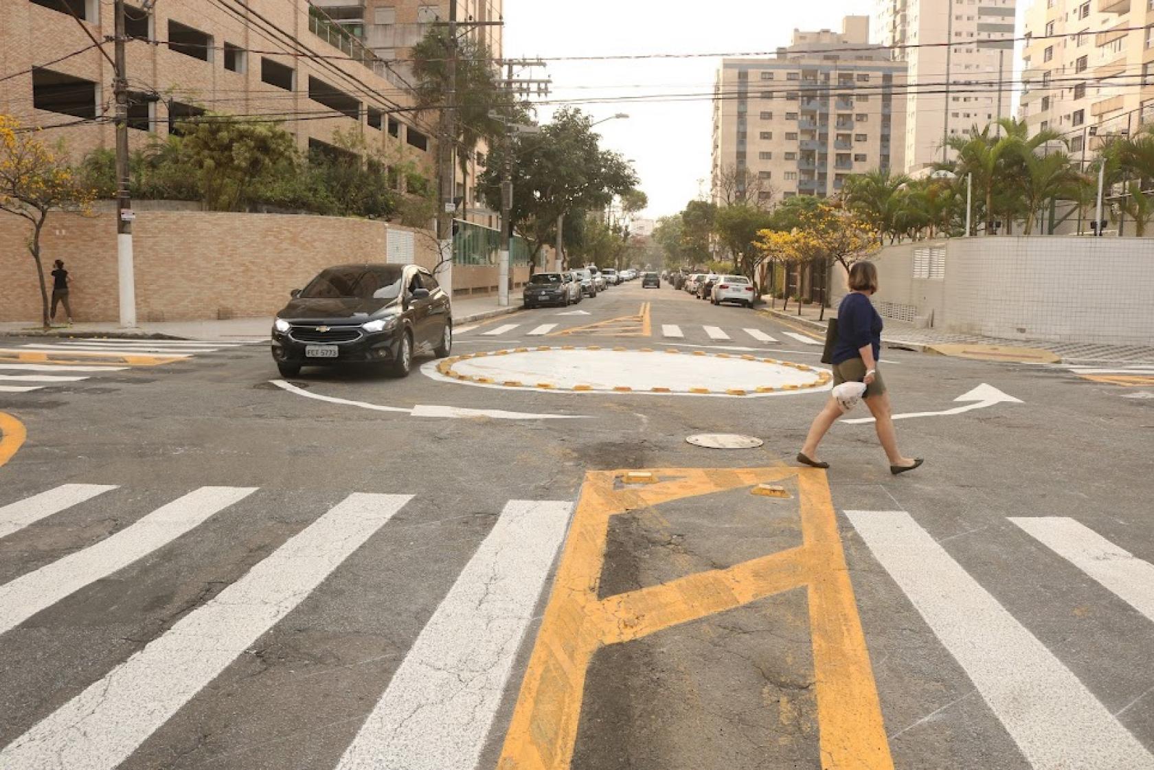 rotatória ao fundo, com carro passando, à esquerda, e mulher atravessando na faixa, à direita. #paratodosverem