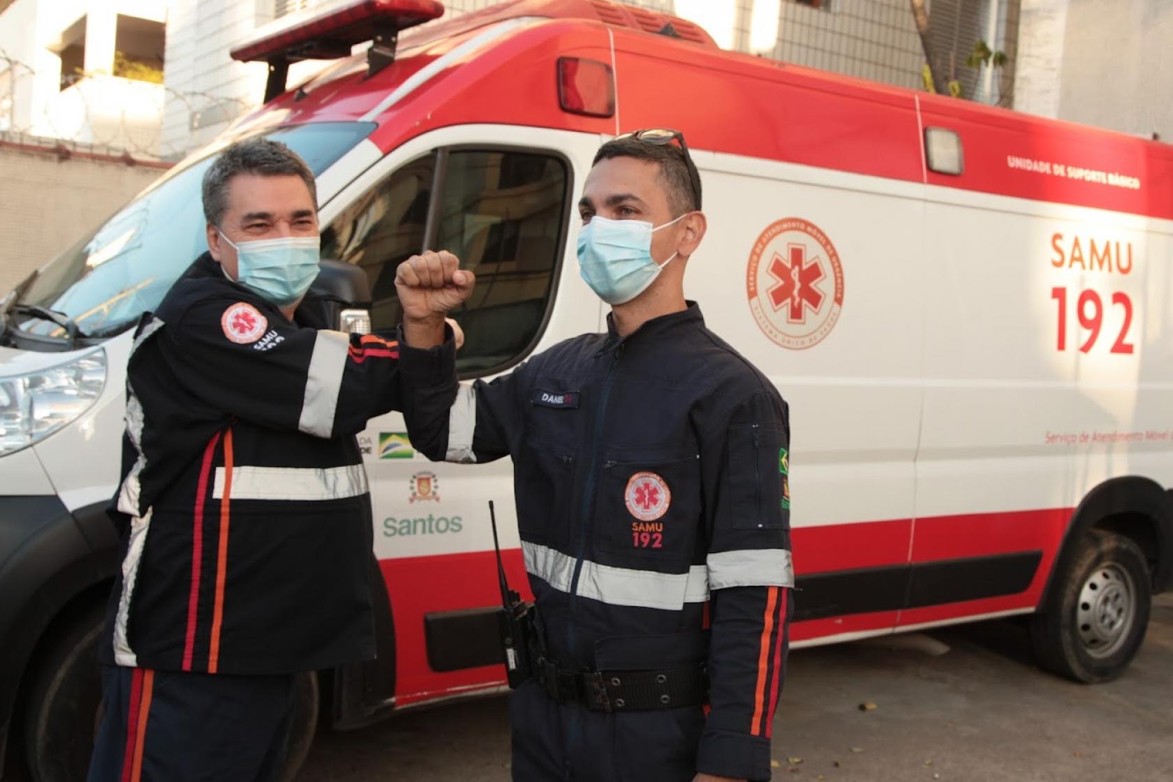 motoristas se cumprimentam tocando os braços. Ao fundo está parada uma ambulância do Suma