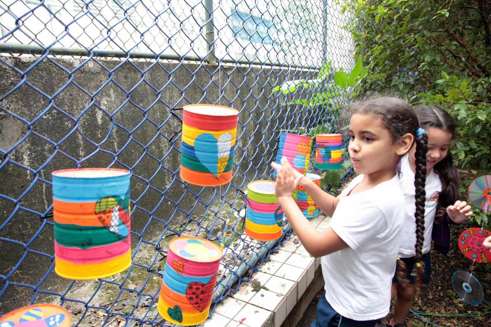 menina brinca com objetos criados com material reciclável, preso em grade de escola #pracegover 
