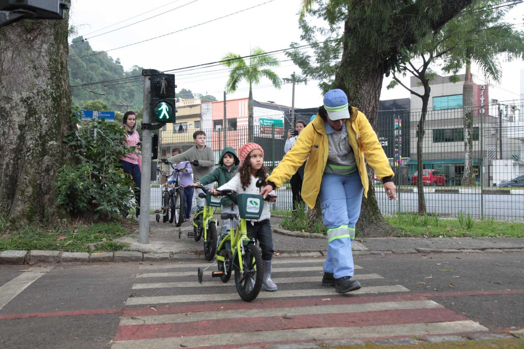 agente ajuda crianças a atravessarem a rua com bicicletas #pracegover 