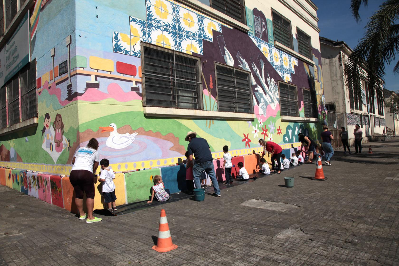 Fachada da escola pintada com vários motivos coloridos. Crianças e professores estão junto à parede finalizando o trabalho. #Pracegover