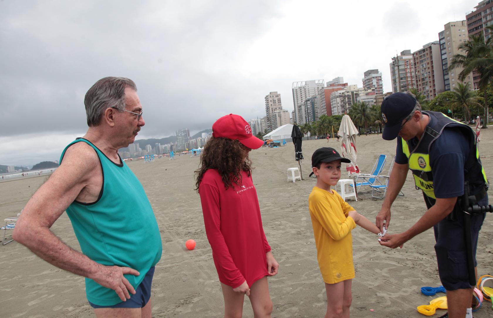 Guarda municipal coloca pulseirinha de identificação em criança na praia. Um adulto e outra criança estão ao lado. #Pracegover