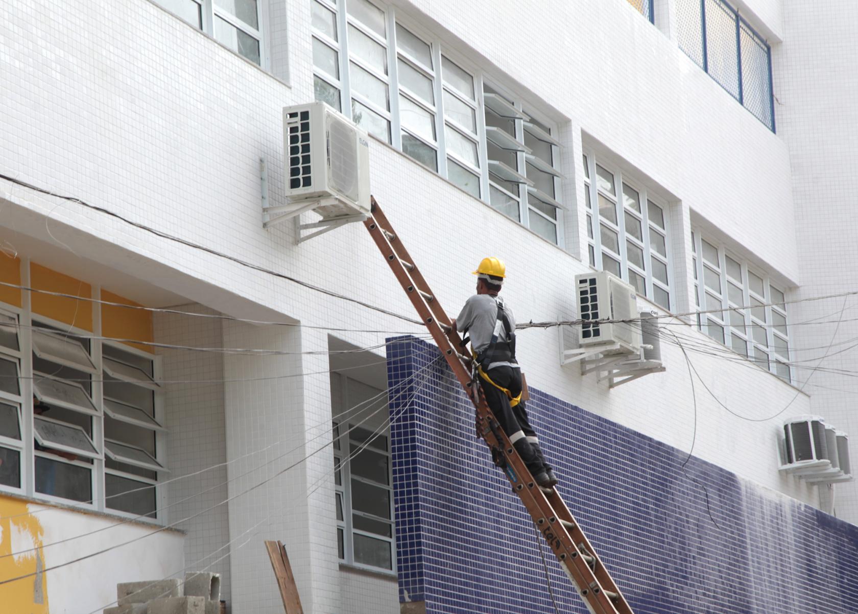 Homem em escada trabalha em parede de escola #pracegover