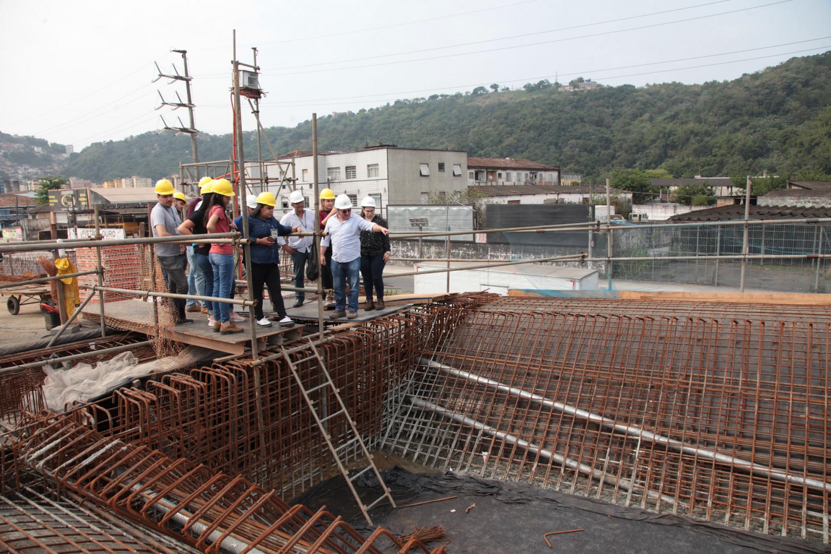 Grupo está observando obra com estruturas de ferro aparentes. Eles estão na altura do solo e olham para baixo, na construção em subsolo. #Pracegover
